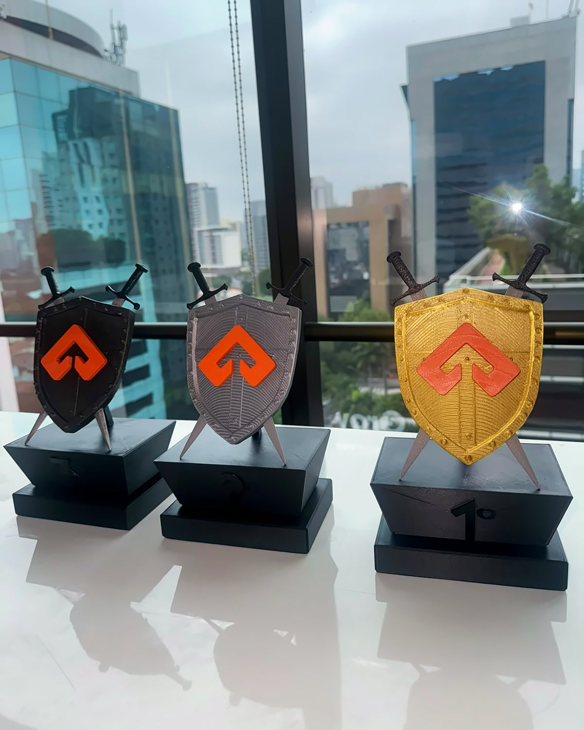 Troféus de 1º, 2º e 3º lugar em formato de escudo com espadas cruzadas, nas cores ouro, prata e bronze, sobre bases pretas